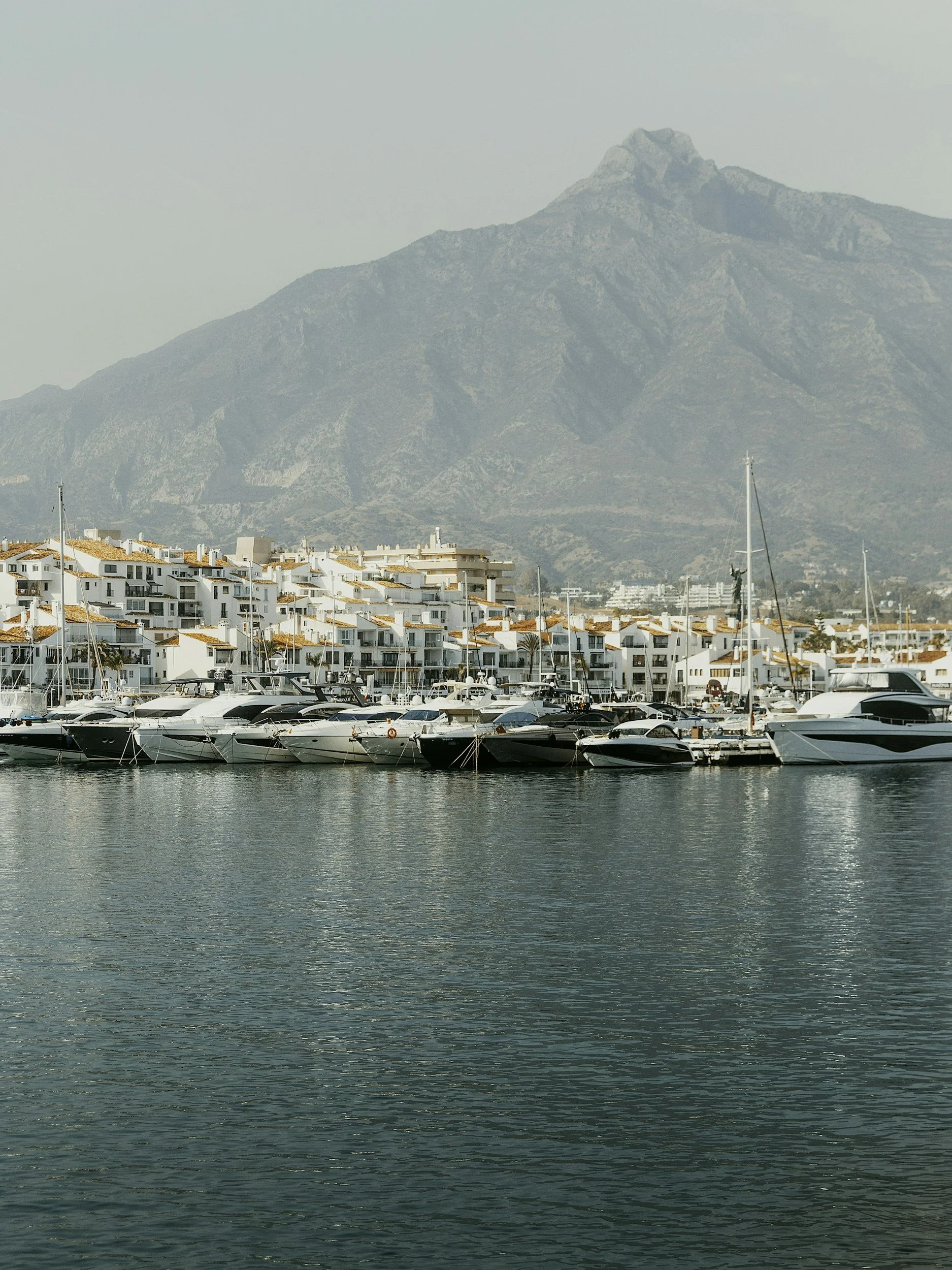 A marina with boats docked on calm water, white buildings with orange roofs in the background, and a large mountain in the distance.