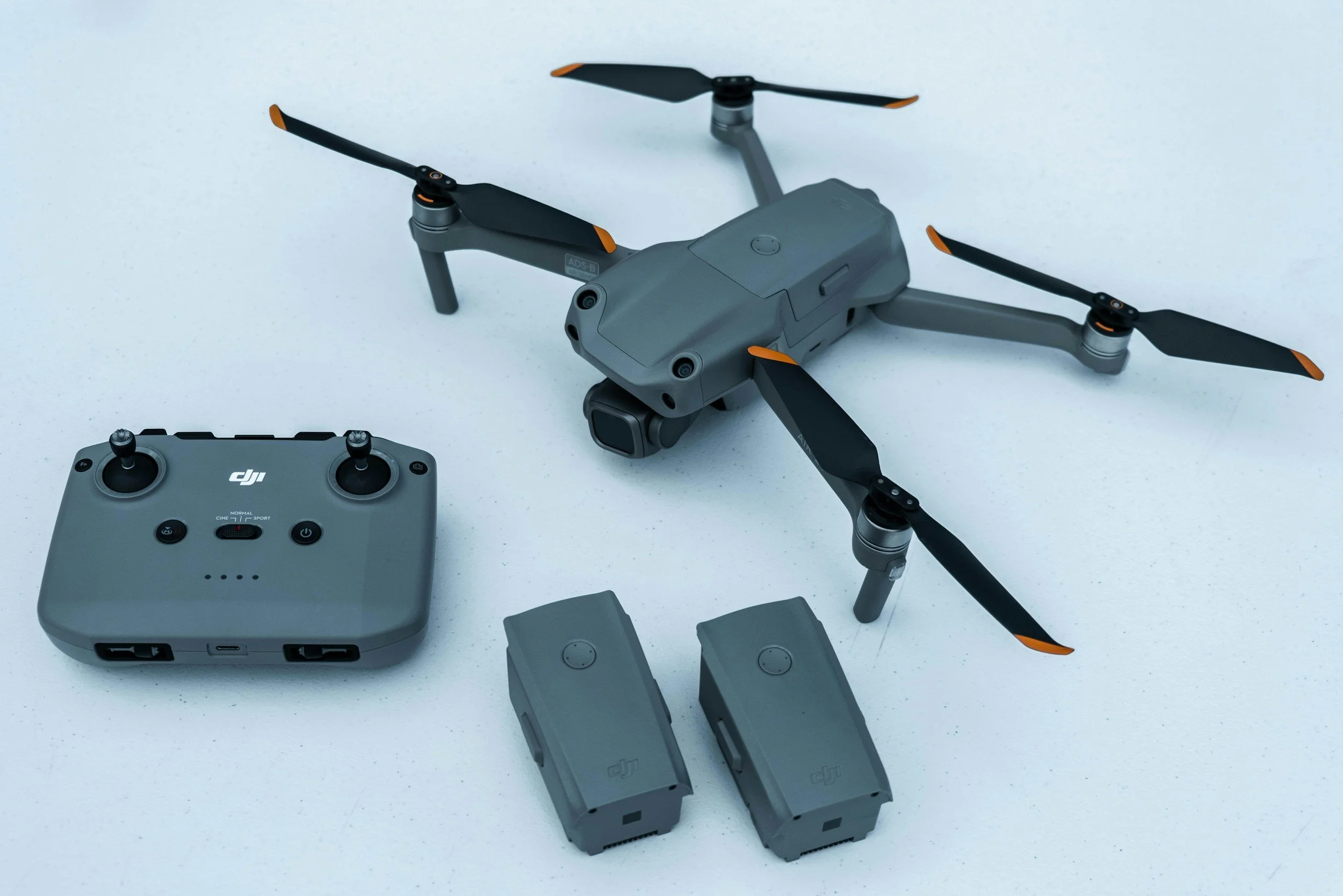 A gray drone with four propellers and orange tips, along with its remote controller and additional batteries, on a white surface.