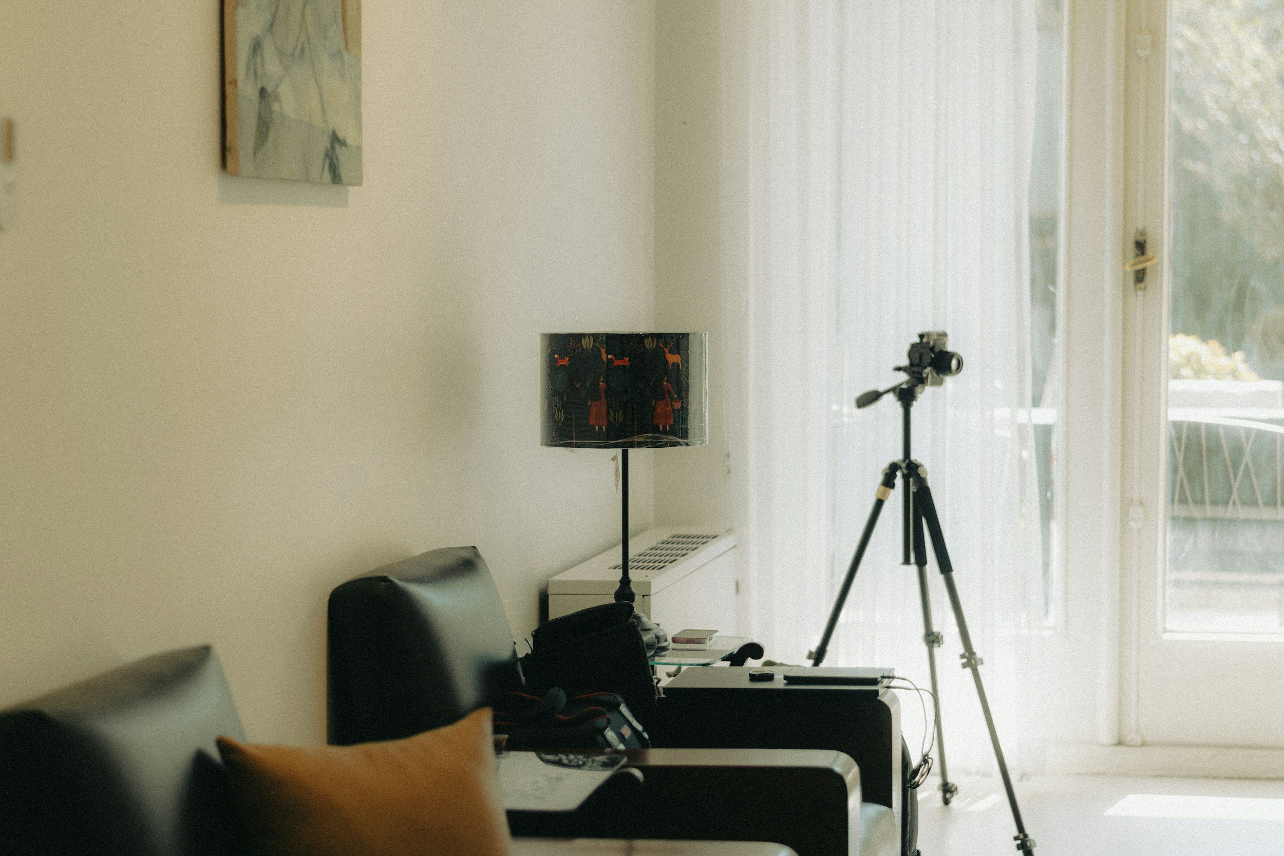 A room with a wall and large windows covered with sheer curtains, a tripod with a camera, a desk with various items, and a lamp with a black shade featuring red and orange designs.
