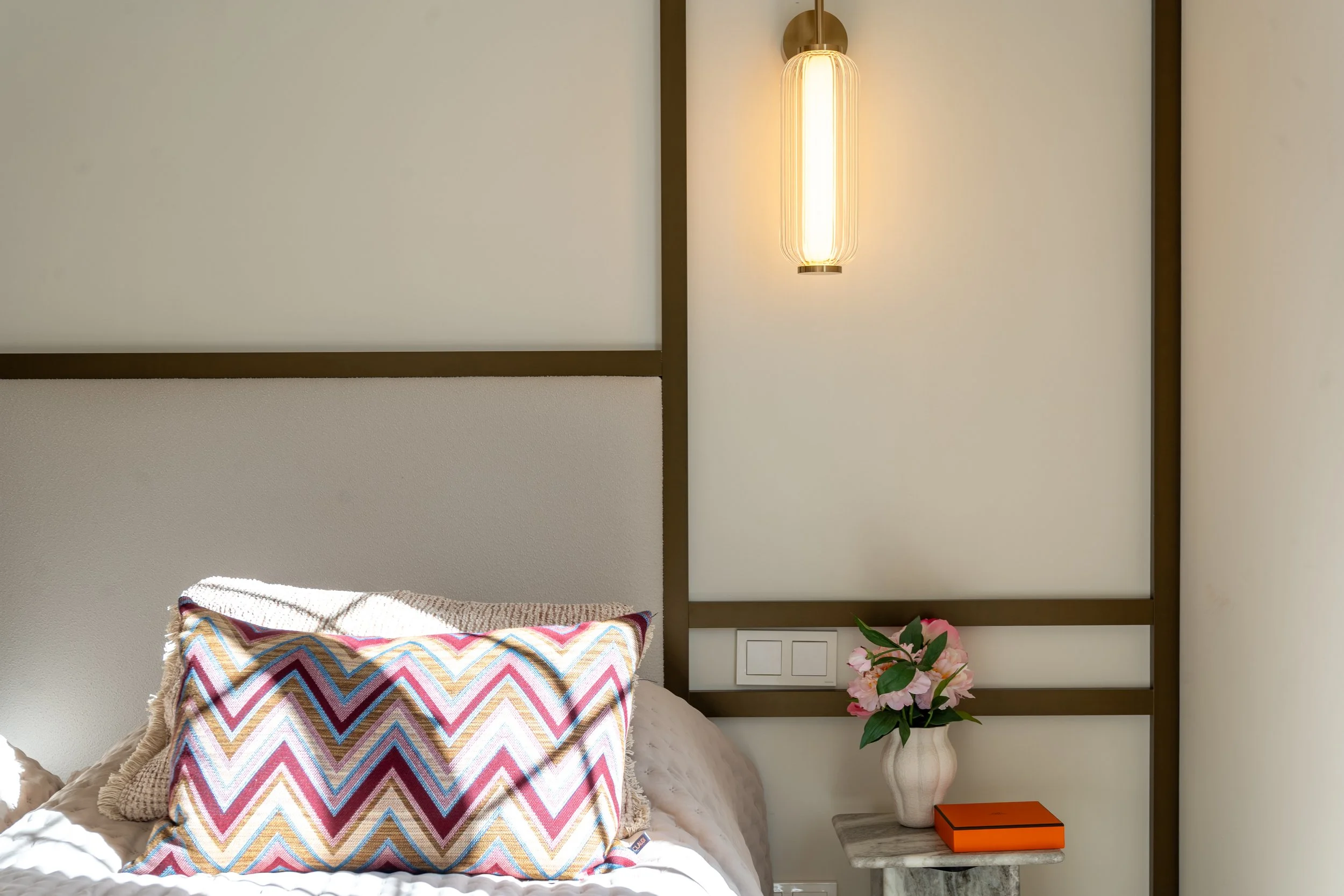 A bedroom wall with a modern hanging light fixture, a pillow with a colorful zigzag pattern, a vase with pink flowers on a small marble table, and part of a bed with cream-colored bedding.