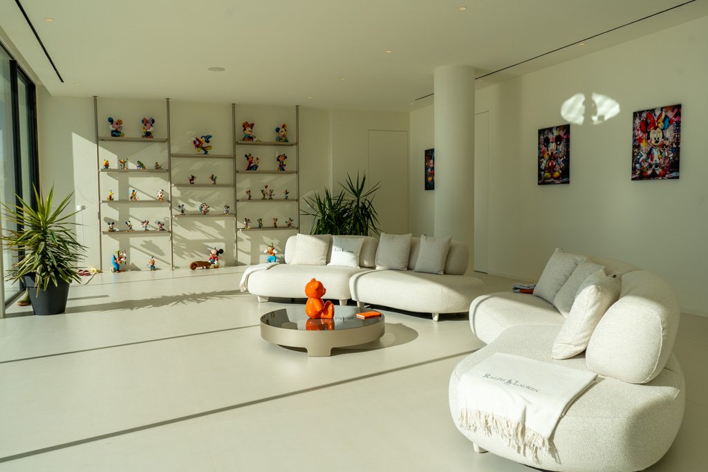 Bright living room with white curved sofas, a coffee table with a small orange toy, book, and blanket, large window with sunlight, shelf with Disney toy figures, and framed Mickey Mouse artwork on wall.