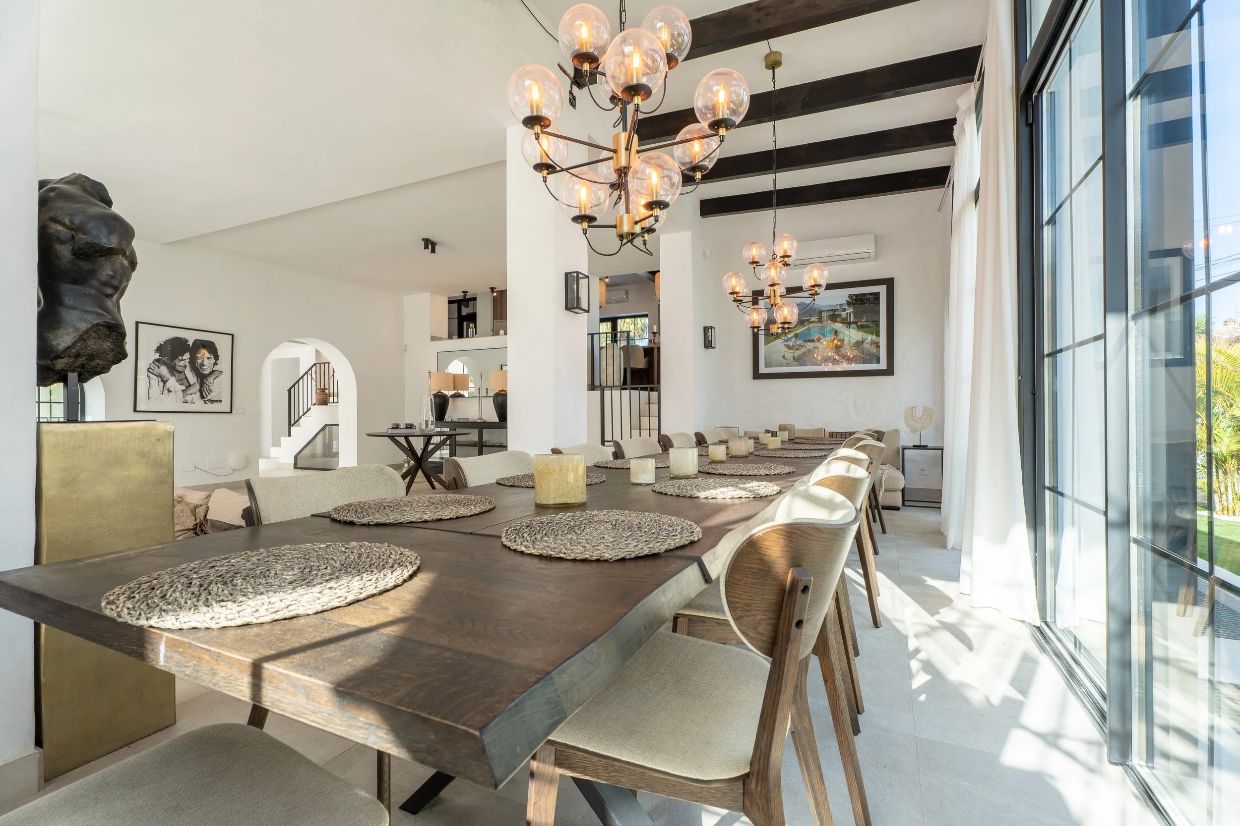 Modern dining room with a long wooden table, woven placemats, and beige chairs. Two chandeliers with multiple bulbs hang from a ceiling with dark wooden beams. Large windows with white curtains allow natural light, highlighting white walls and contemporary artwork.