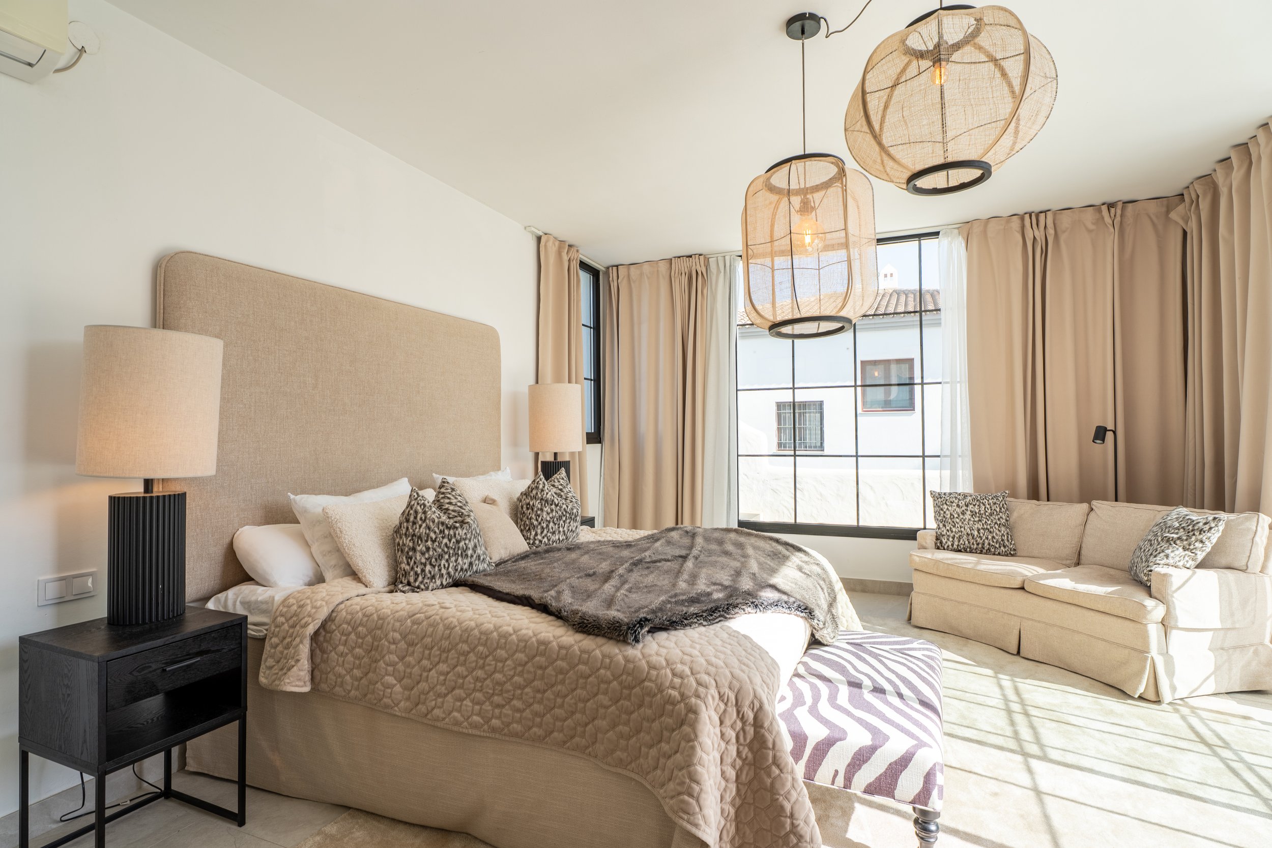 A modern bedroom with a beige upholstered bed, black side table, beige curtains, and pendant lights hanging from the ceiling near large windows showing a building outside.