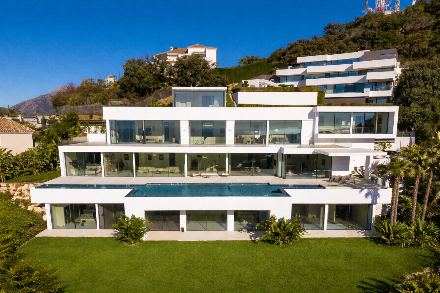Modern white multi-level house on a hillside with large glass windows, a rooftop pool, and lush landscaping.