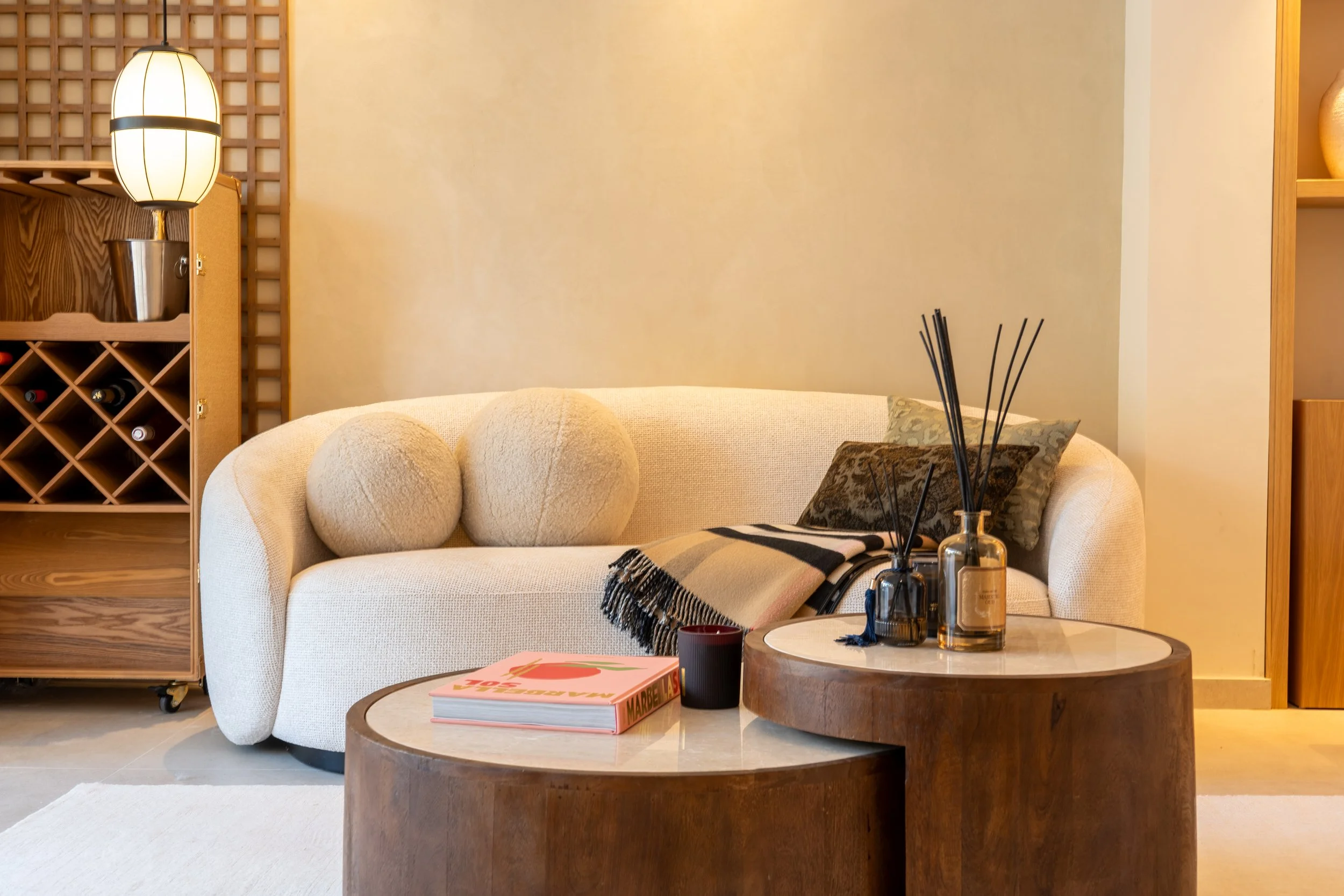 Living room with beige sofa, black and beige pillows, wooden coffee table with books and decorative items, a lamp, and wooden shelves with wine bottles and a decorative vase.