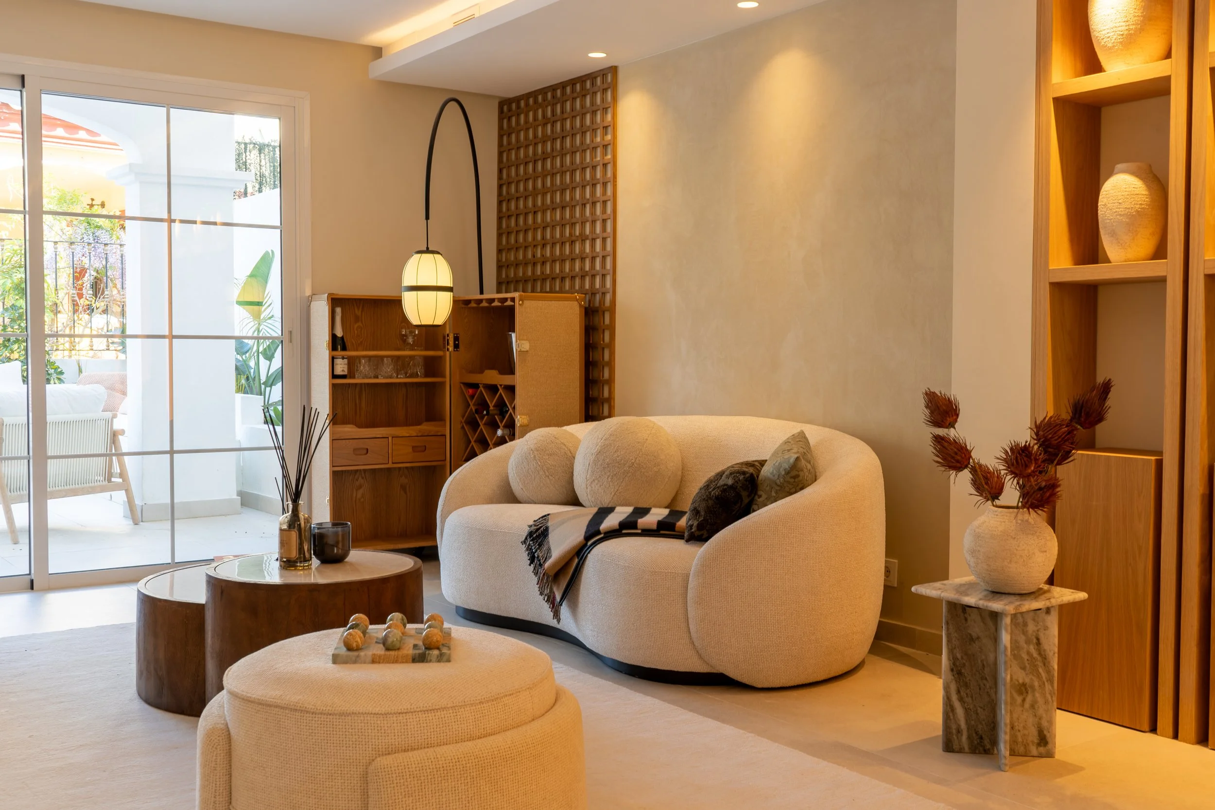 A cozy living room with a beige sofa, round ottoman, and wooden coffee table. There's a decorative vase with dried flowers, a room lamp, and a sliding glass door leading to a balcony with outdoor furniture and plants.