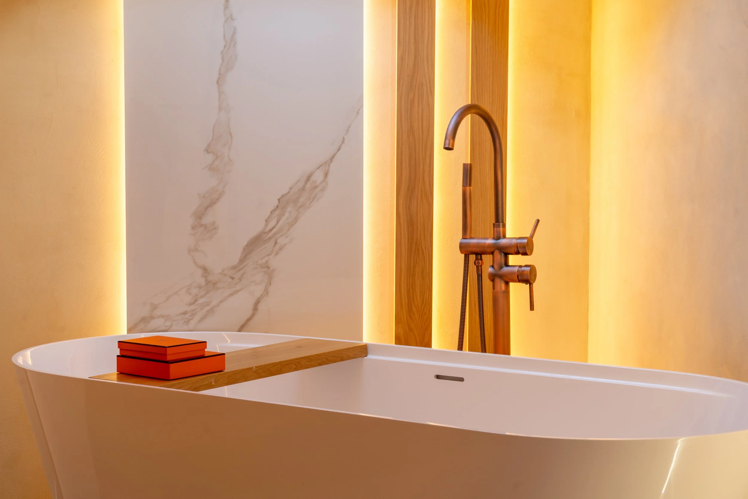 Modern bathroom sink with a copper faucet, a stacked set of orange books on the edge, and a marble wall with vertical wood accents and soft lighting.