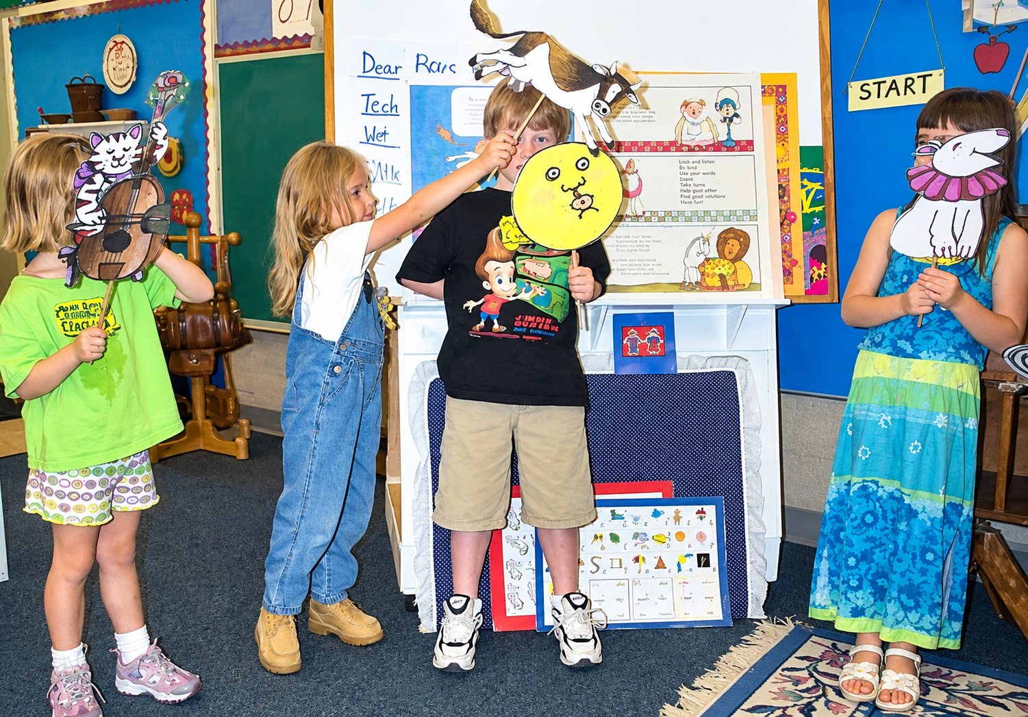 Five children in a classroom holding puppets; a girl in a neon green shirt and patterned shorts, a girl in a white shirt and denim overalls, a boy in a black T-shirt and khaki shorts, and two girls with masks of cartoon characters.