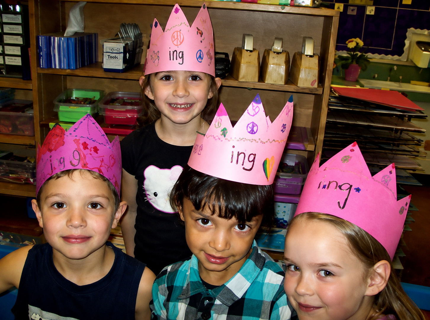 Group of four children wearing pink paper crowns decorated with various drawings and the word 'ing', celebrating in a classroom setting.
