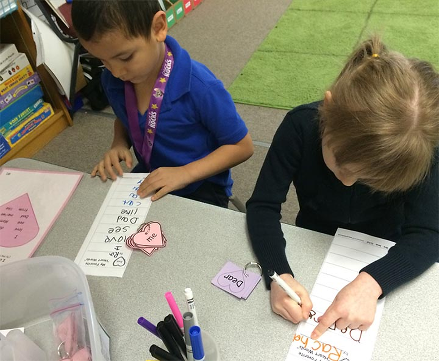 Two children sitting at a classroom table working on a writing activity. One child is writing on a sheet with the word "Race" written on it, while the other child is pointing at their paper with a marker. There are pink and purple heart-shaped flashcards on the table with the word "me" and "Dear" written on them.