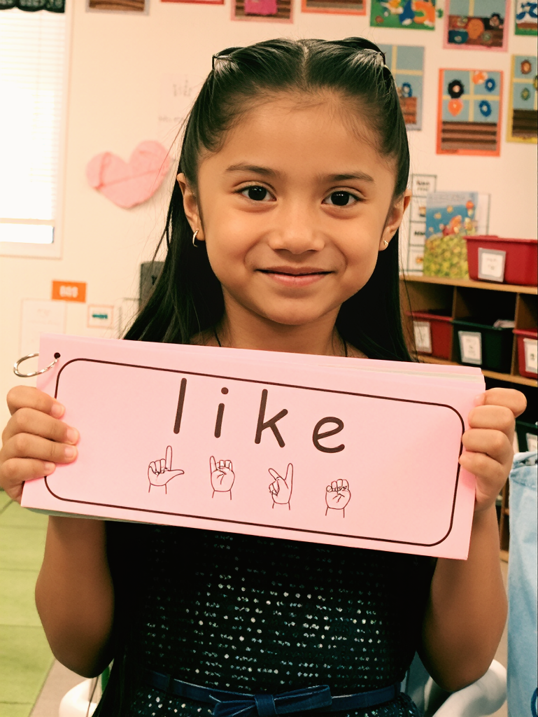 A young girl with long dark hair, smiling, holding a pink sign that says "like" with American Sign Language fingerspelling for 'like' below each letter, in a classroom with colorful decorations.