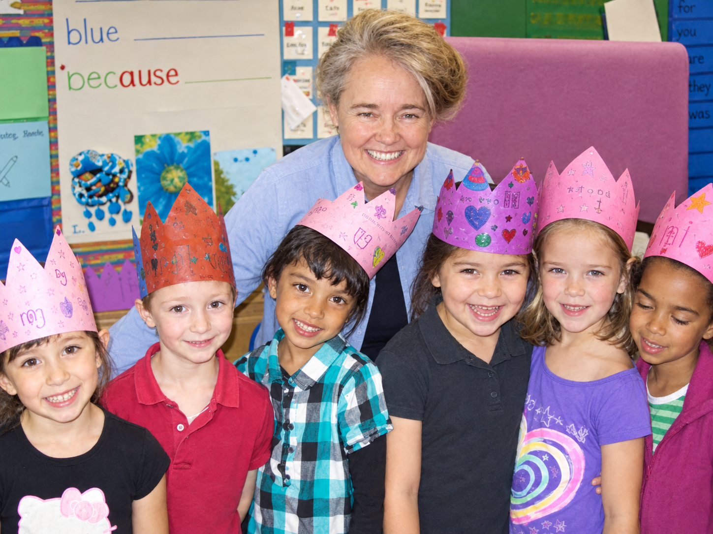 A teacher and six young children wearing colorful paper crowns, smiling, in a classroom with educational posters and bright decorations.