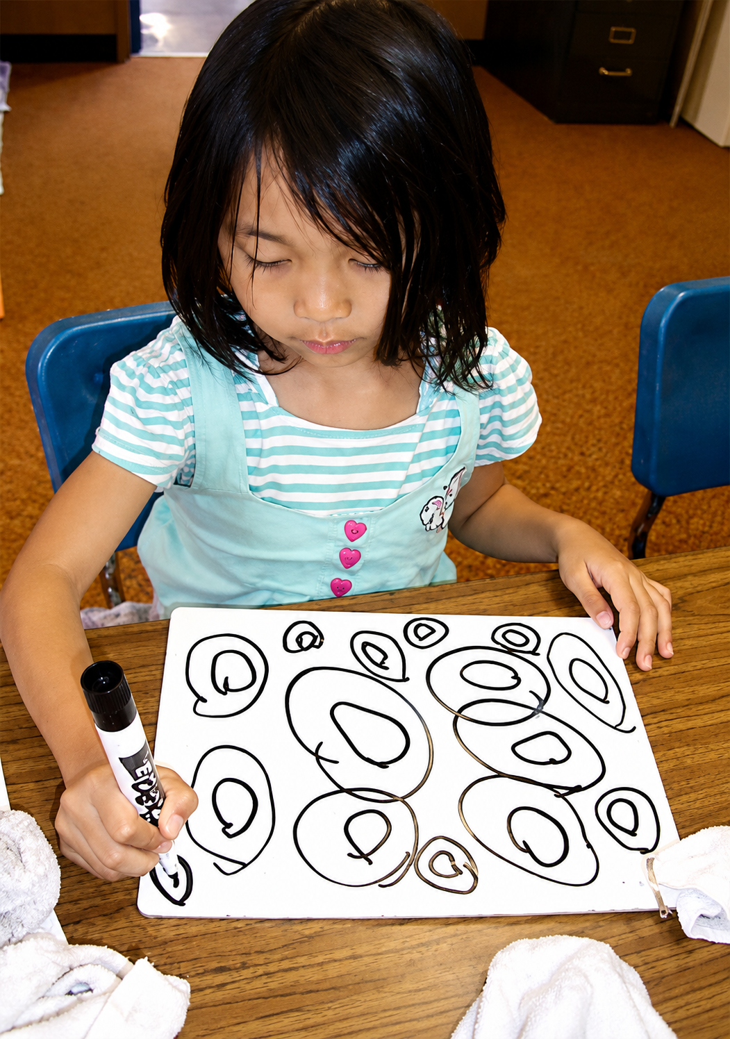 A young girl with wet hair drawing black circles on a white paper with a marker at a wooden table.