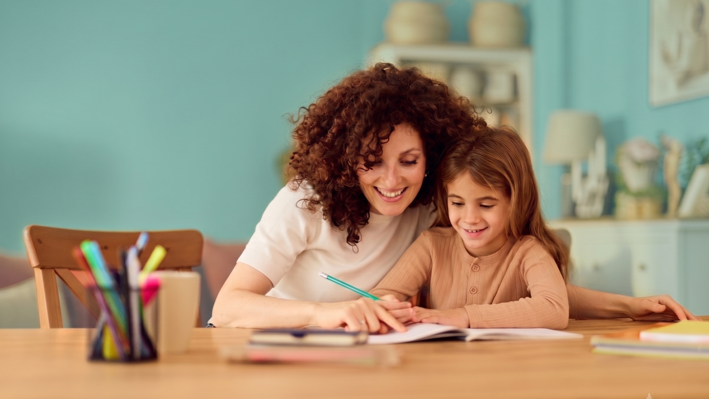A woman and a young girl sit at a wooden table, smiling and writing on paper together in a brightly decorated room.