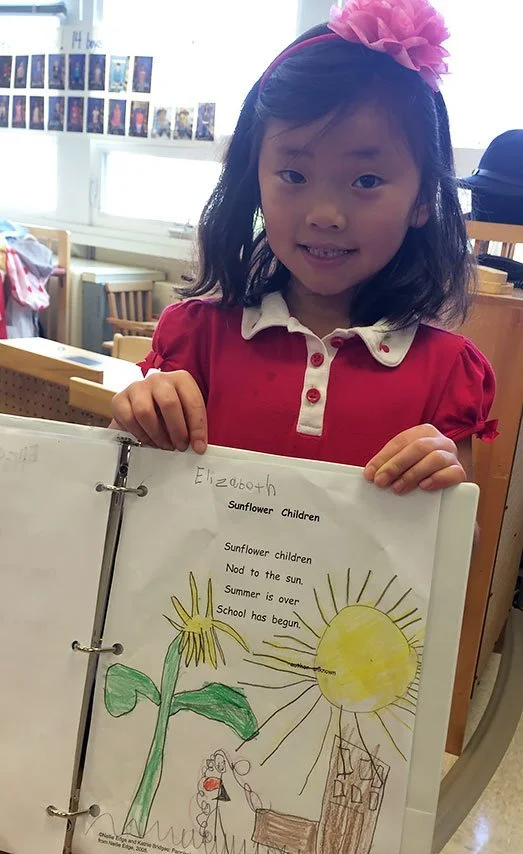 A young girl with a pink headband and red dress holding a notebook with a drawing of a sunflower and a poem about sunflower children in a classroom.