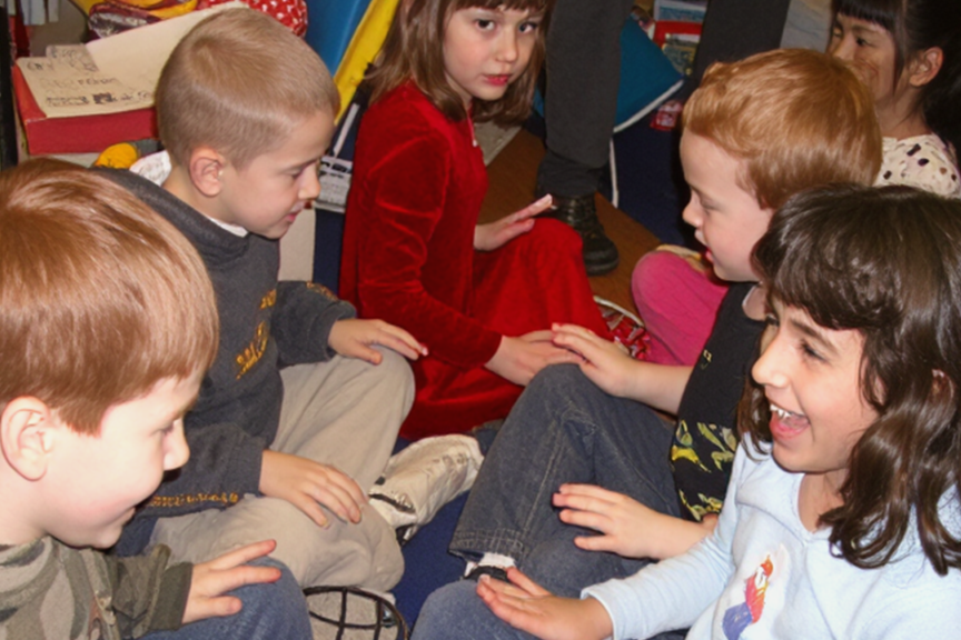 Children sitting on the floor, engaged in a group activity, with a girl in a white shirt smiling and interacting with a boy in a black shirt.