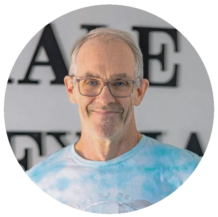 A middle-aged man with short gray hair, glasses, and light facial hair smiling at the camera. He is wearing a tie-dye blue and white shirt. In the background, there is a wall with partially visible large black letters.