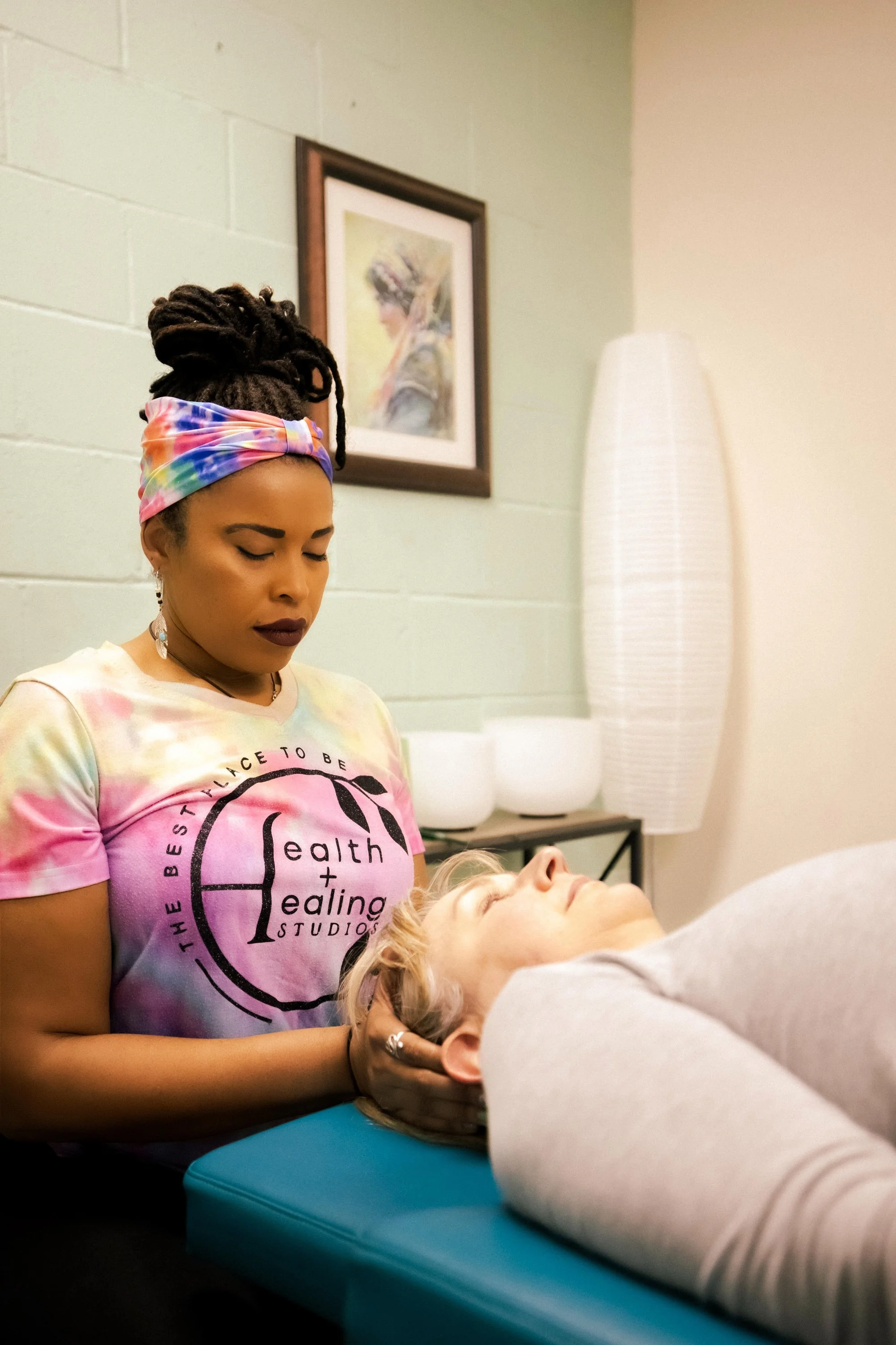 A woman with dreadlocks and a colorful headband is giving a treatment to a person lying on a treatment table, with her eyes closed, in a wellness or therapy room. The woman is wearing a tie-dye t-shirt with the text 'the best I like to be' and 'health + healing studios', and there is a framed picture on the wall and a tall lamp in the background.