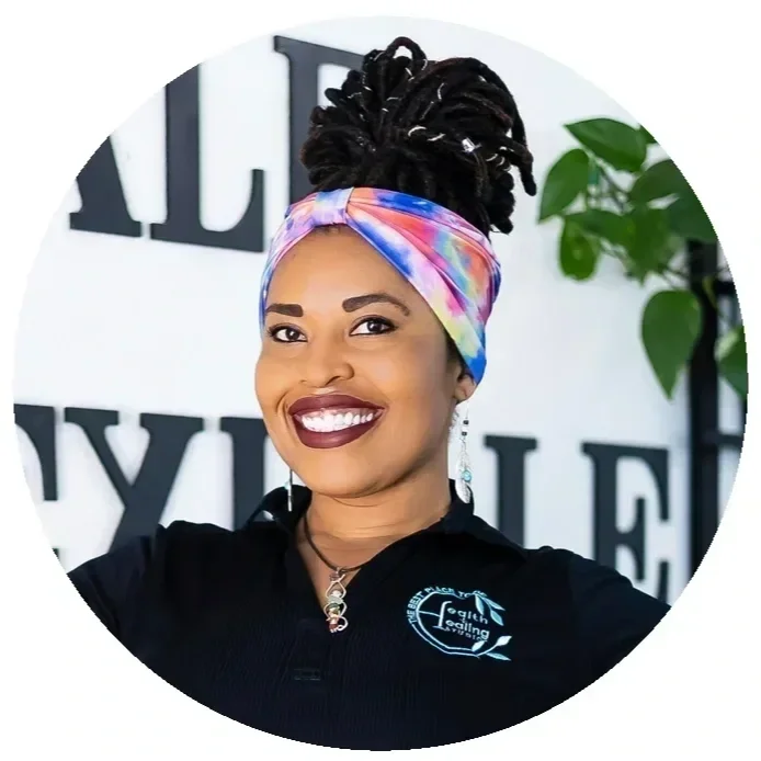 A smiling Black woman with dark lipstick and earrings, wearing a colorful headscarf and a black shirt with a logo, standing indoors with white walls and large black text in the background, next to a green potted plant.