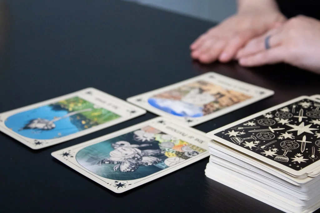 A person's hands resting on a black table near a spread of tarot cards and a large deck of cards with black and white celestial designs.