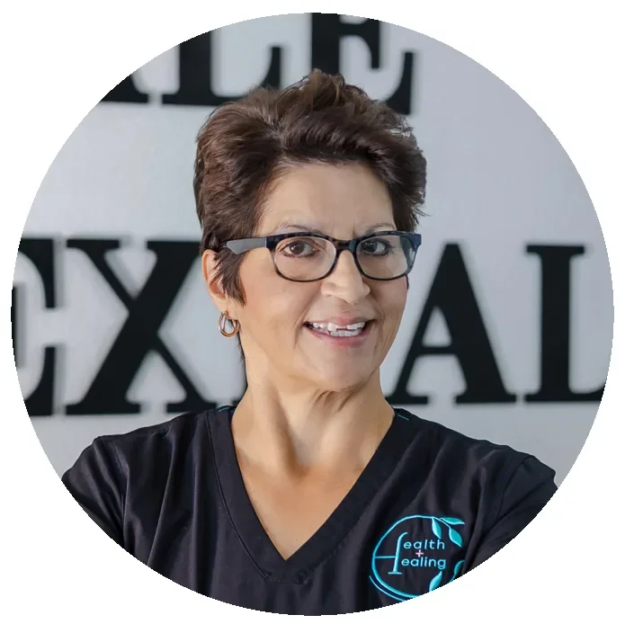 A woman with short dark hair, glasses, and earrings, smiling, wearing a black shirt with a logo that says "Health & Healing". She is standing in front of a background with black and white letters.