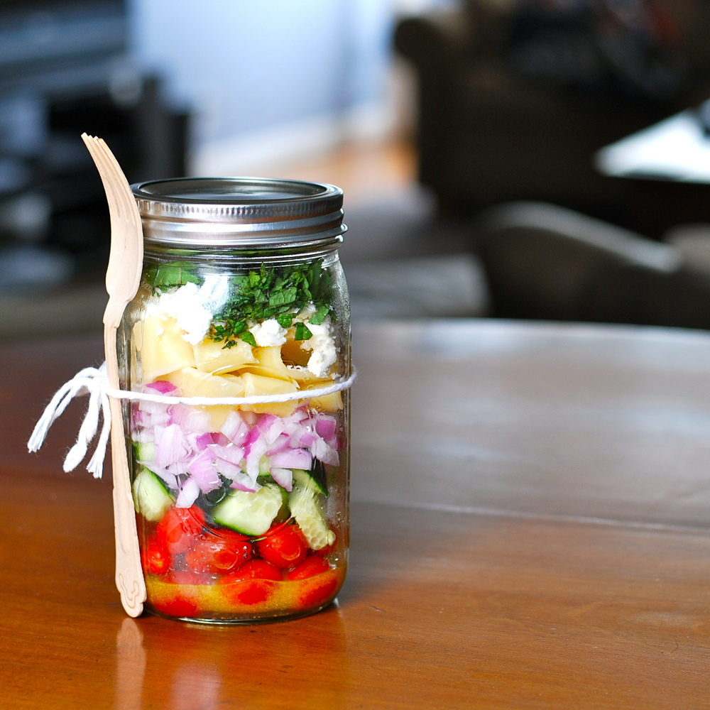 A glass jar filled with layered fresh vegetables including cherry tomatoes, cucumbers, onions, cheese, and herbs. A wooden fork is attached to the jar with a string, placed on a wooden table.