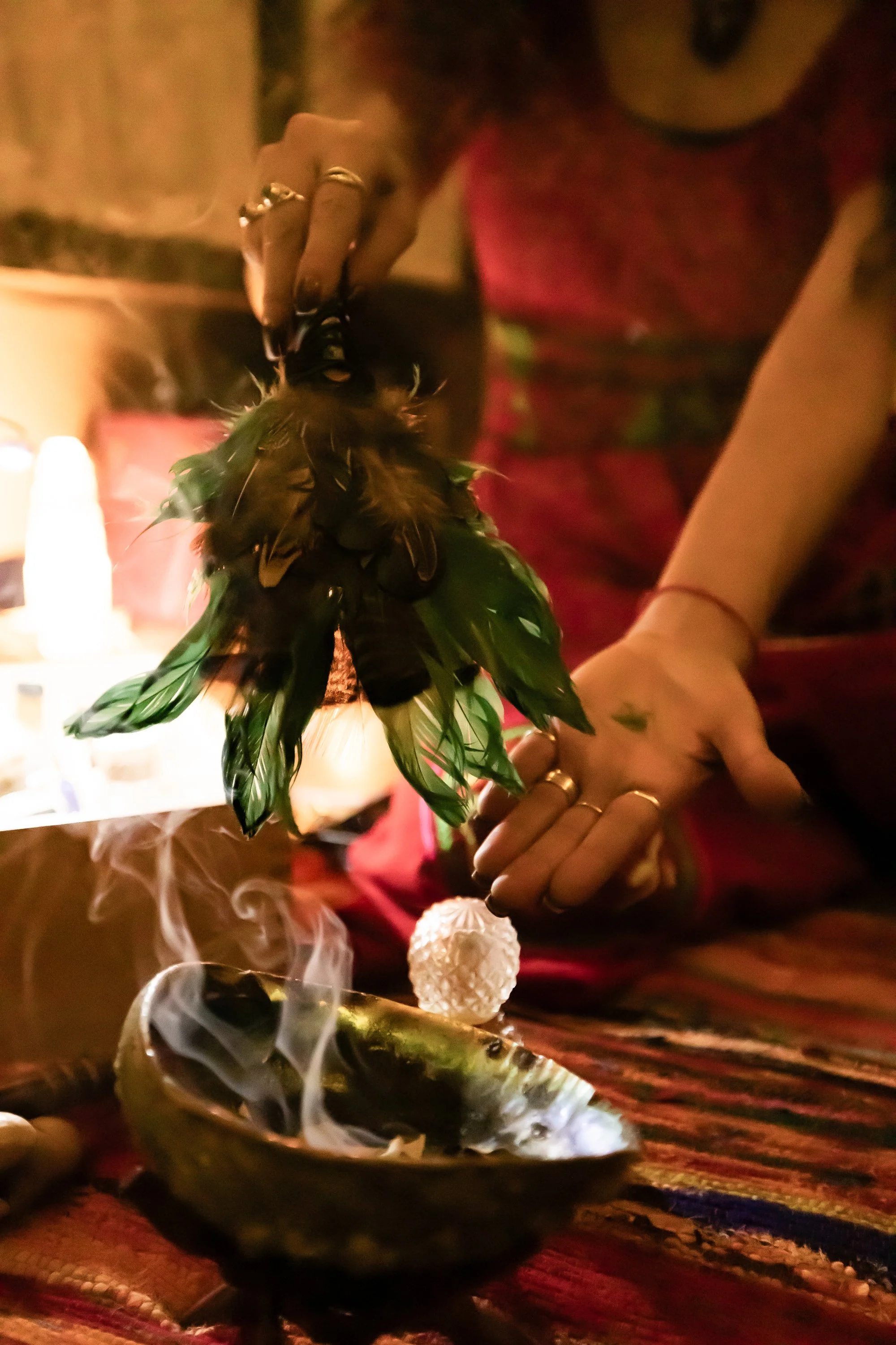A person with rings on their fingers is holding a feathered object over a small fire with smoke rising from it, in a dimly lit setting.