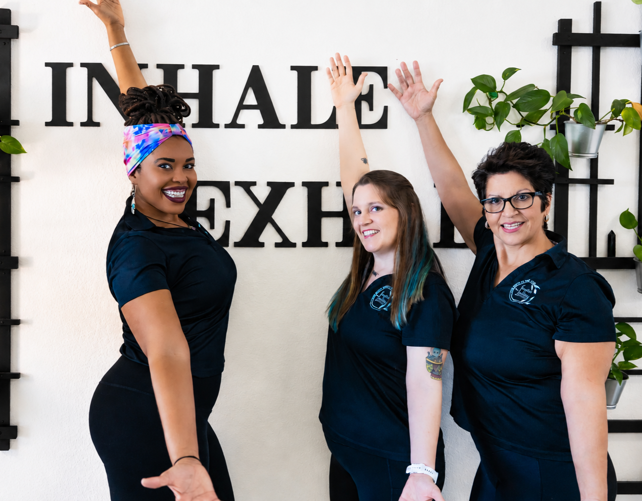 Three women in black shirts standing in front of a wall with the word 'INHALE' in large black letters, smiling and raising hands. One woman has a colorful headscarf, another has blue-tinted hair, and the third has short dark hair and glasses.