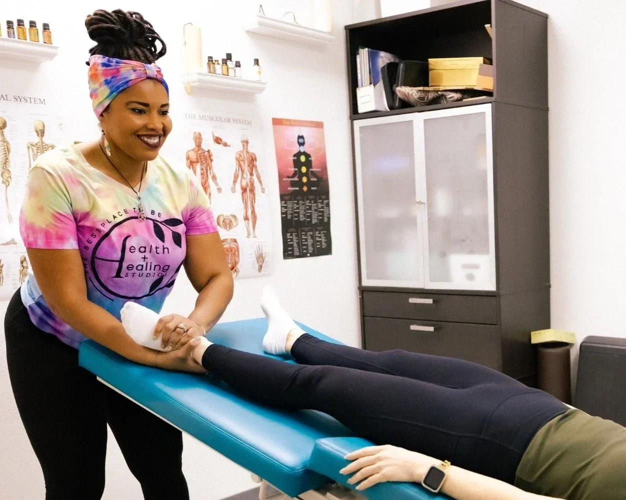 A woman with a colorful headscarf and a rainbow tie-dye shirt providing therapy or treatment to a patient lying face down on a medical table in a clinical setting with anatomical posters on the wall and medical supplies on a cabinet.