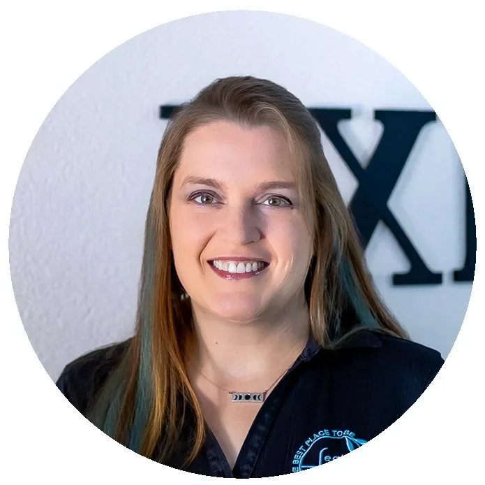 A woman with long light brown hair, smiling, wearing a black shirt with a logo, in front of a white wall with large black letters.