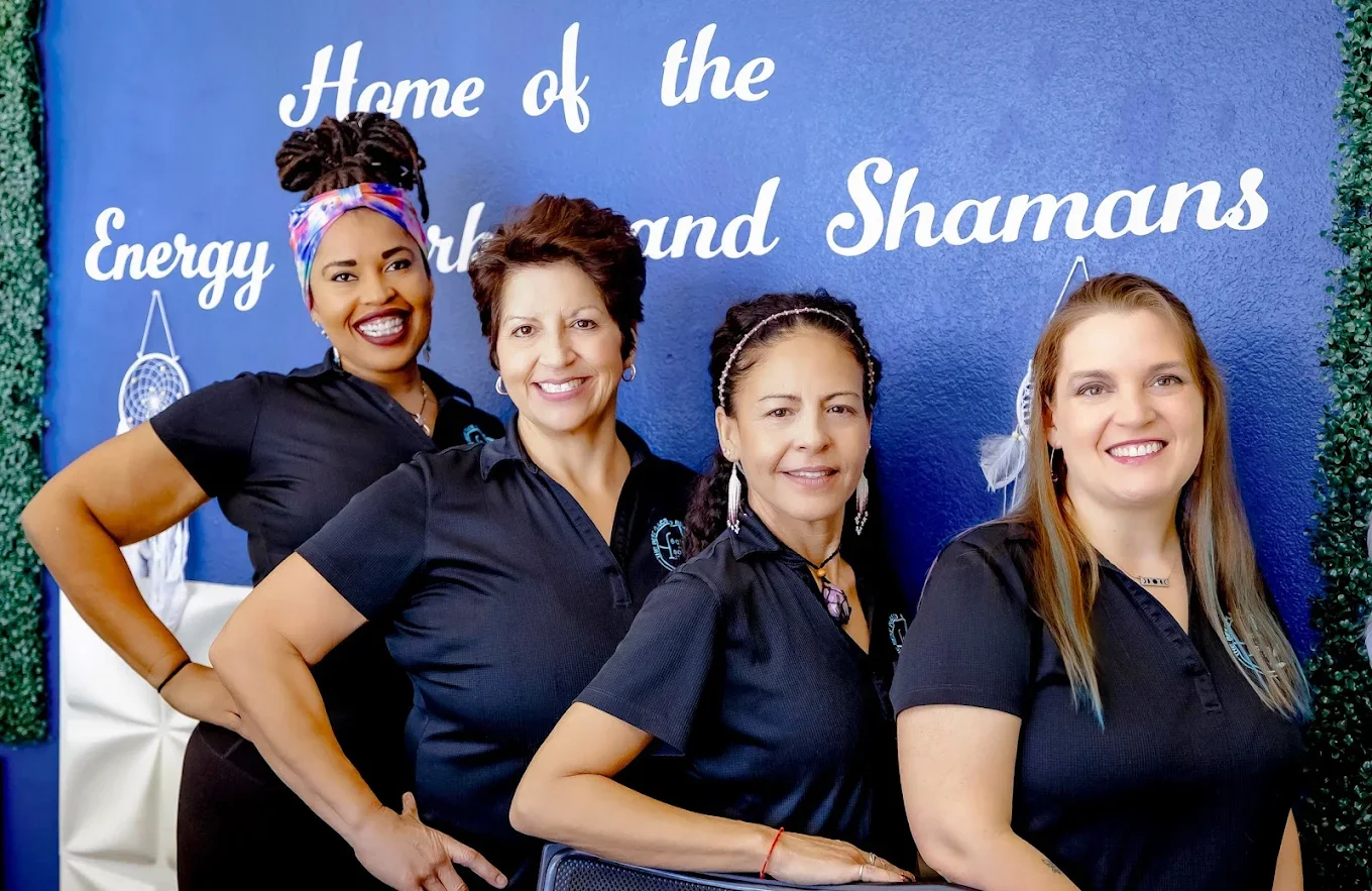 Four women standing in front of a blue wall with white text that reads 'Home of the Energy Heartland and Shamans.' They are all wearing black shirts and smiling.