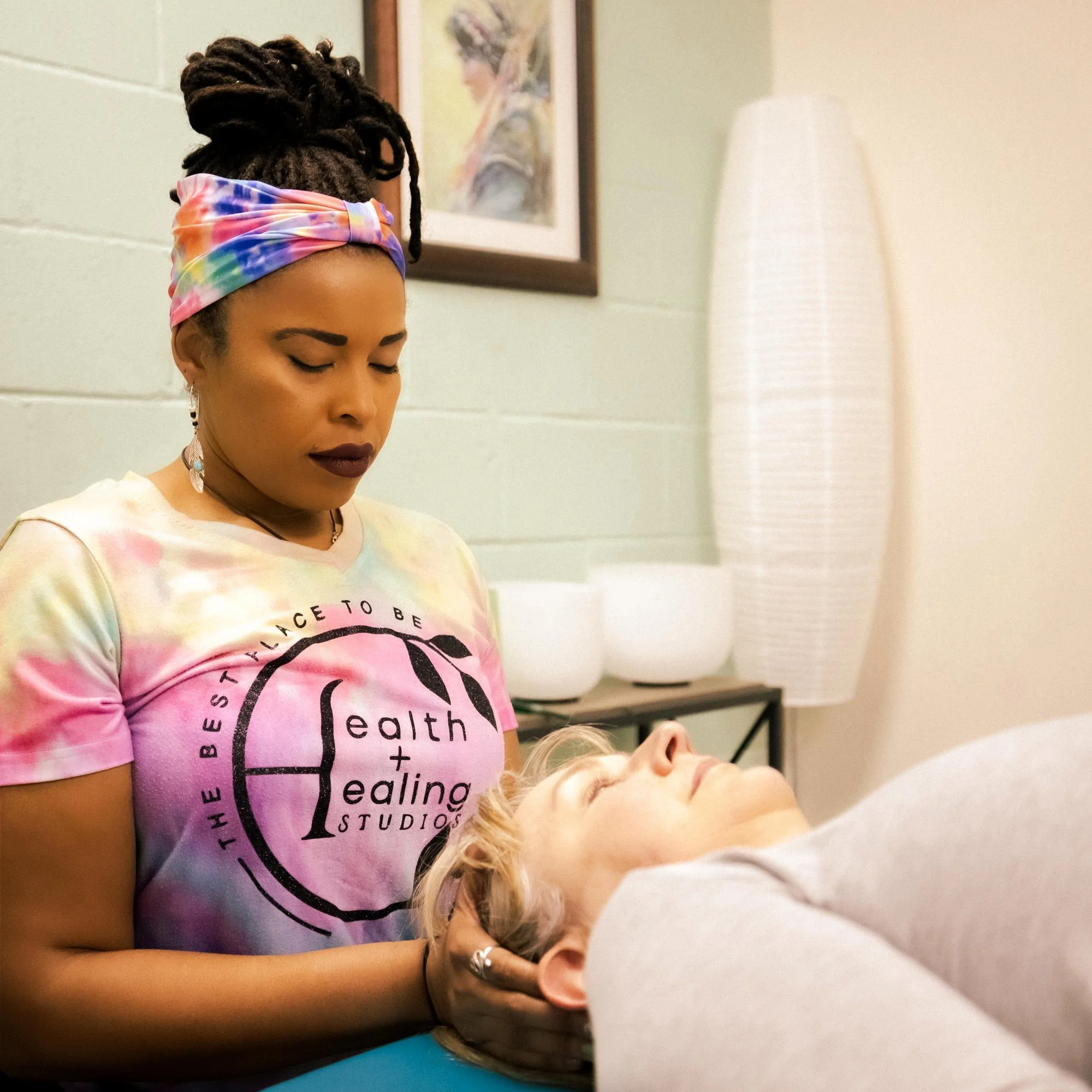A woman with dreadlocks wearing a colorful tie-dye headband and a tie-dye shirt with the words 'healing, strength, healing' printed on it, is performing a procedure on a person's forehead in a clinical or studio setting.