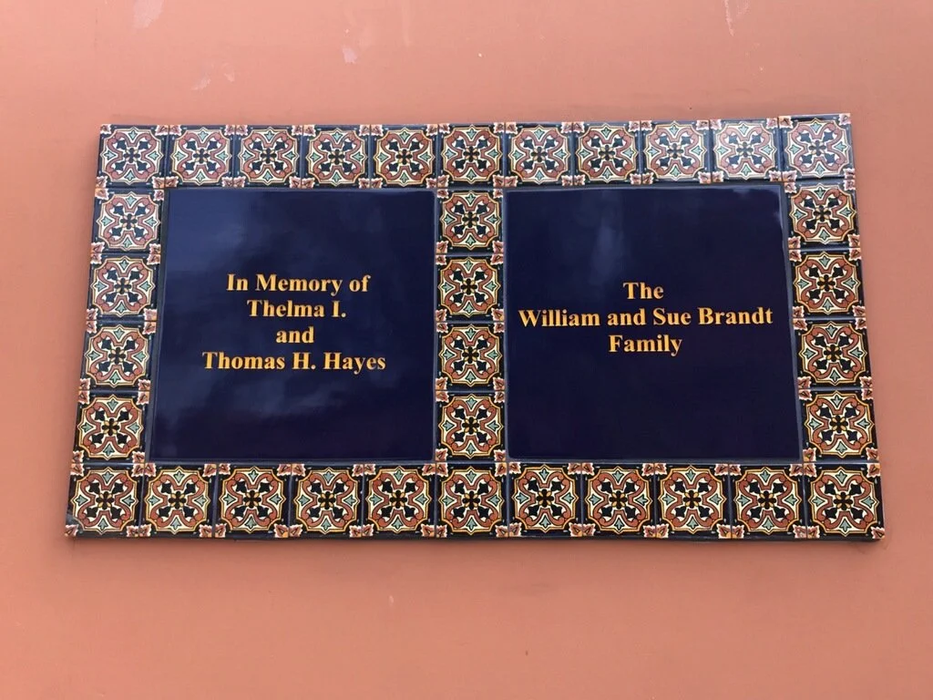Two tiles with blue backgrounds and gold text are surrounded by a decorative border of patterned tiles. The left tile reads 'In Memory of Thelma I. and Thomas H. Hayes,' and the right tile reads 'The William and Sue Brandt Family.'