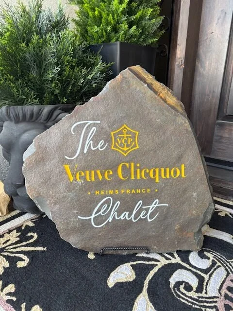 A large decorative stone sign with the text 'The Veuve Clicquot Reims France Chalet' engraved and painted in white and yellow, placed outdoors near potted plants and a wooden fence.