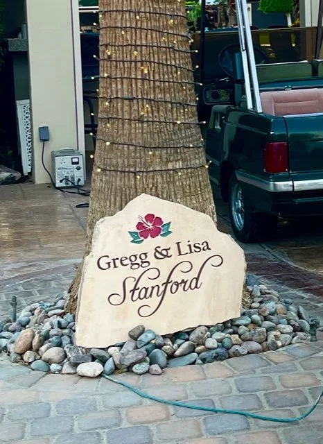 A large stone sign with the names Gregg and Lisa Stanford, topped with a pink flower and green leaves, placed near a tree wrapped with string lights in an outdoor setting.