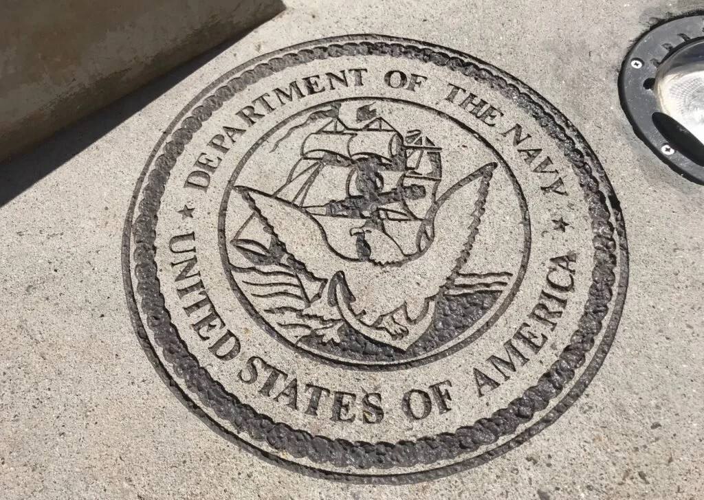 Seal of the United States Department of the Navy on concrete sidewalk, featuring a ship, eagle, and banner.