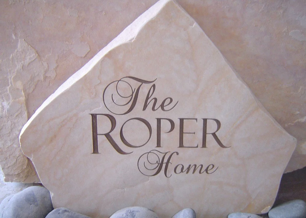 A stone sign reading 'The Roper Home' in decorative font, placed among small rocks.