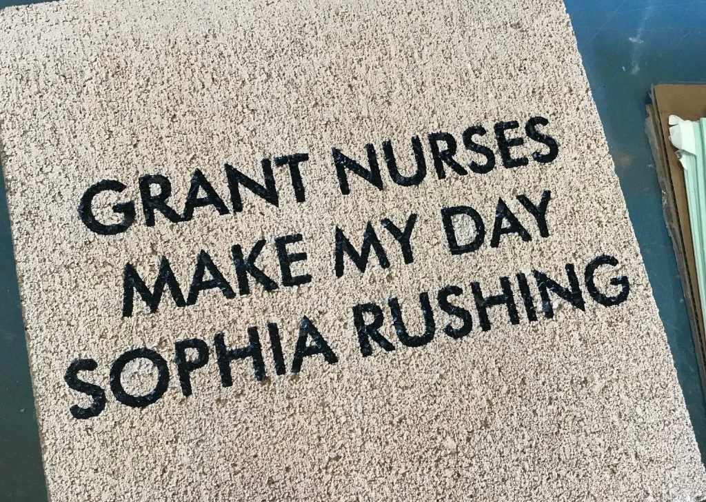 A beige doormat with black text that reads: 'Grant Nurses Make My Day Sophia Rushing'.