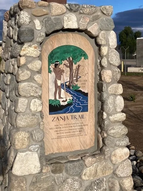 A stone monument with a wooden plaque depicting a Native American man standing near a stream, trees, and a river, with the text 'Zanja Trail' and a description of its historical significance.