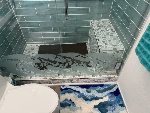 View of a glass-enclosed shower area with gray and white pebble tile accents, a shower bench, and a dark drain mat; part of a bathroom with a white toilet and a colorful blue, white, and beige patterned rug.