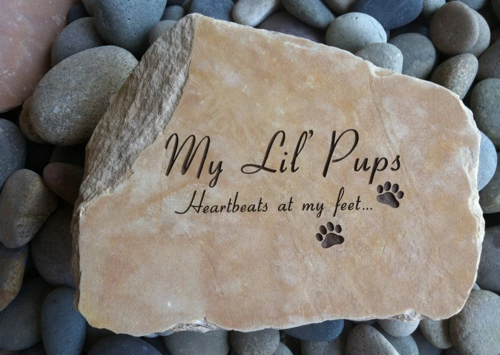 A decorative stone engraved with the words "My Lil Pups Heartbeats at my feet..." and paw prints, placed on a bed of smooth pebbles.