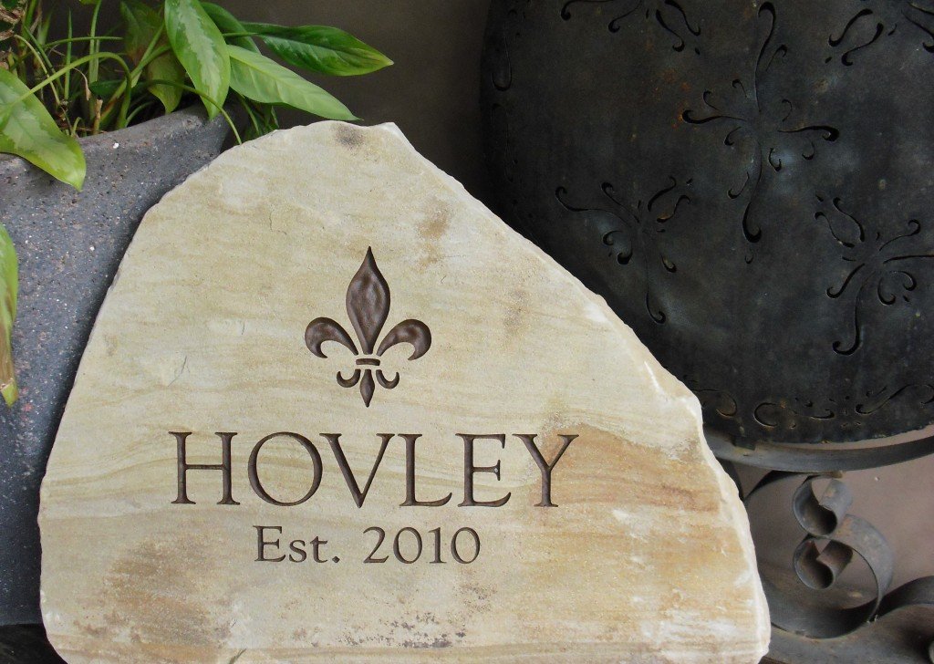 A decorative stone with the name 'Hovley' and the text 'Est. 2010,' featuring a fleur-de-lis symbol above the name, surrounded by potted plants and garden ornaments.