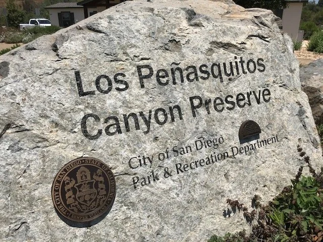 A large rock with engraved text that reads 'Los Peñasquitos Canyon Preserve,' along with the City of San Diego Park & Recreation Department seal and a small dark opening in the rock.