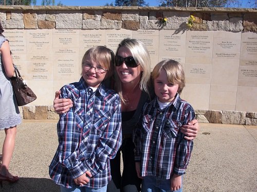 A woman with blonde hair and sunglasses smiling, hugging two young boys wearing plaid shirts, standing in front of a stone wall with engraved plaques.