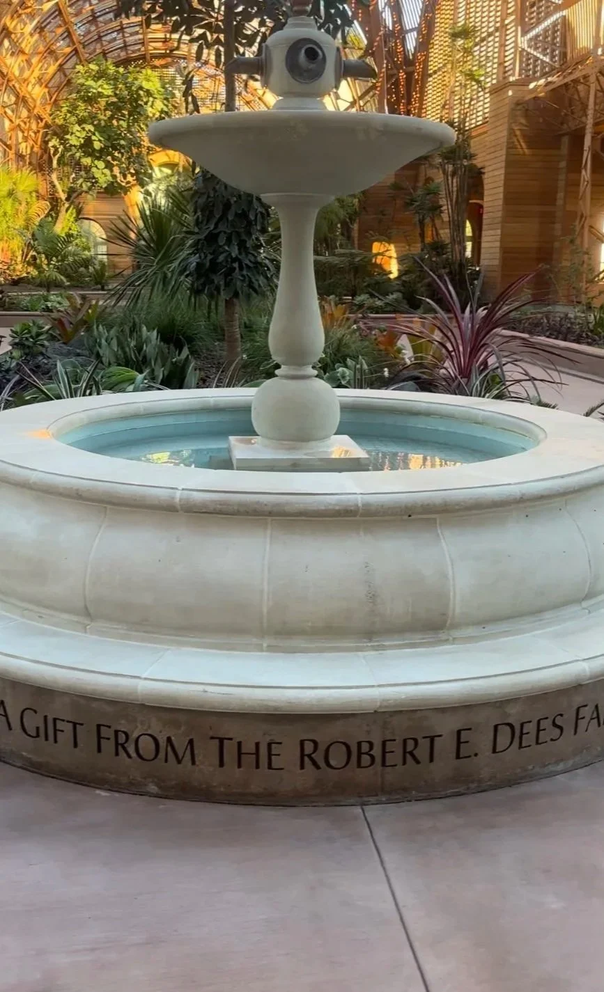 A decorative fountain with a satellite-shaped camera on top, surrounded by plants and garden in an indoor botanical garden with glass ceiling.