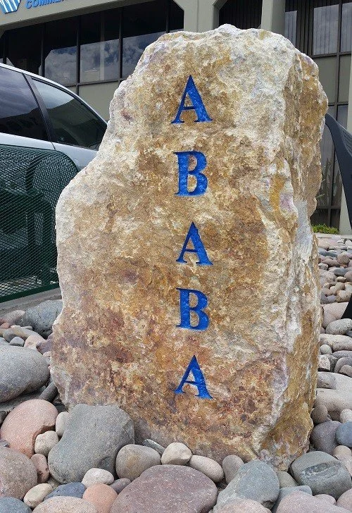 Large beige-colored rock with the word "ABBA" written vertically in blue capital letters, placed among smaller rocks outside a building.