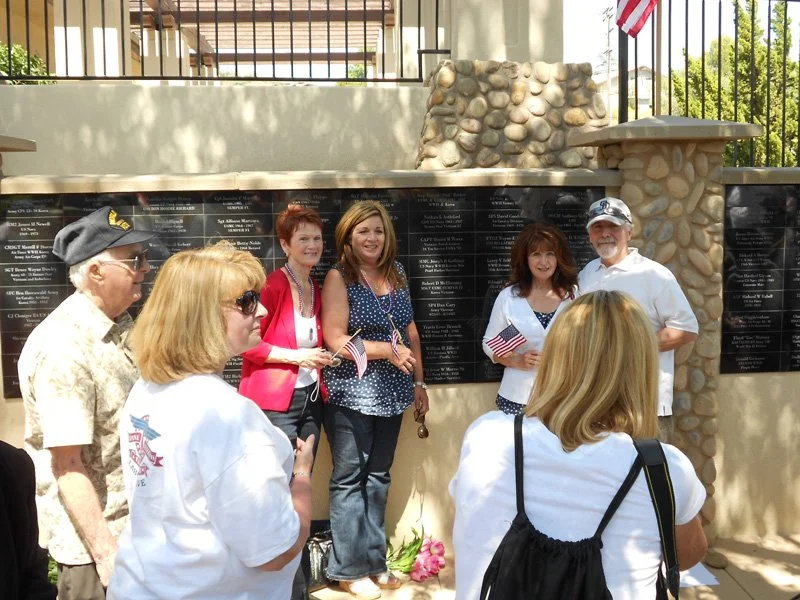 Group of people gathered at a memorial wall with plaques, some holding small American flags, at a sunny outdoor location with an American flag in the background.