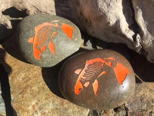 Two rocks with fish-shaped carvings painted in orange, placed outdoors among other stones.