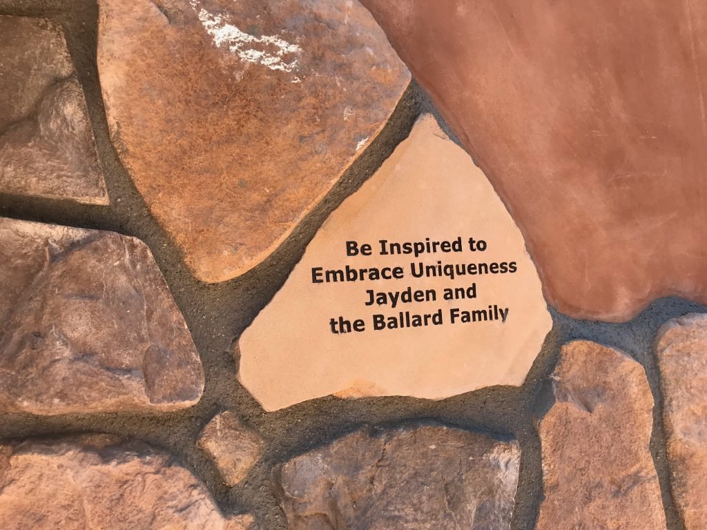 A stone wall with a beige stone engraved with the inspirational message: 'Be Inspired to Embrace Uniqueness Jayden and the Ballard Family'.