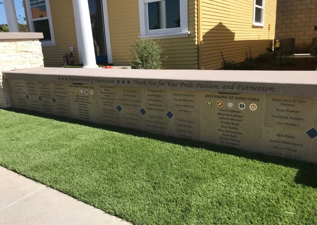 A concrete wall with engraved text and emblems honoring board of directors, set in front of a yellow house with a window and door, with neatly trimmed grass in the foreground.